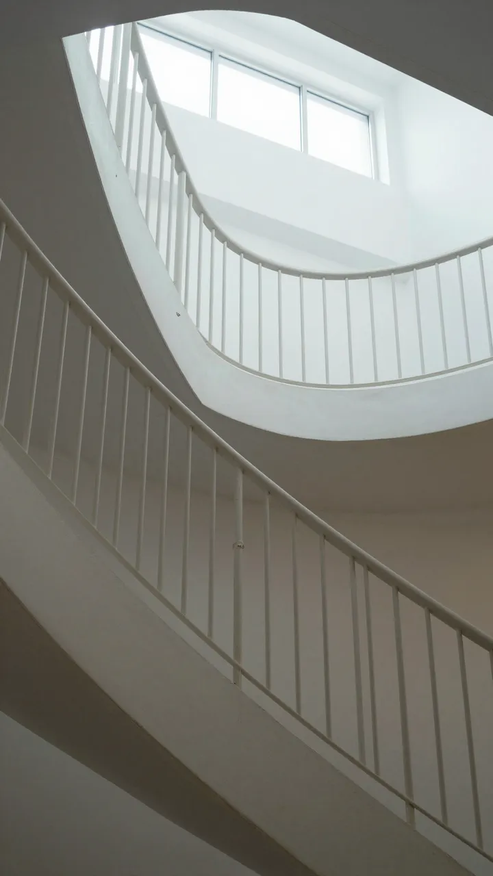 Vertical view up a curved white staircase with railings and overhead natural daylight from a window