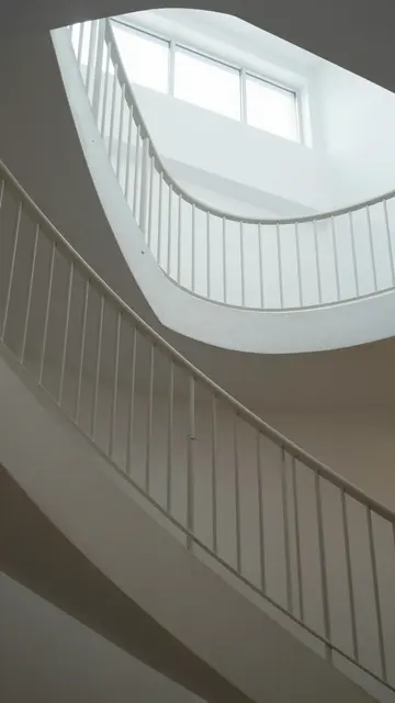 Vertical view up a curved white staircase with railings and overhead natural daylight from a window