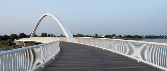 View along a curving modern white pedestrian bridge over a calm river at dusk, with no people present