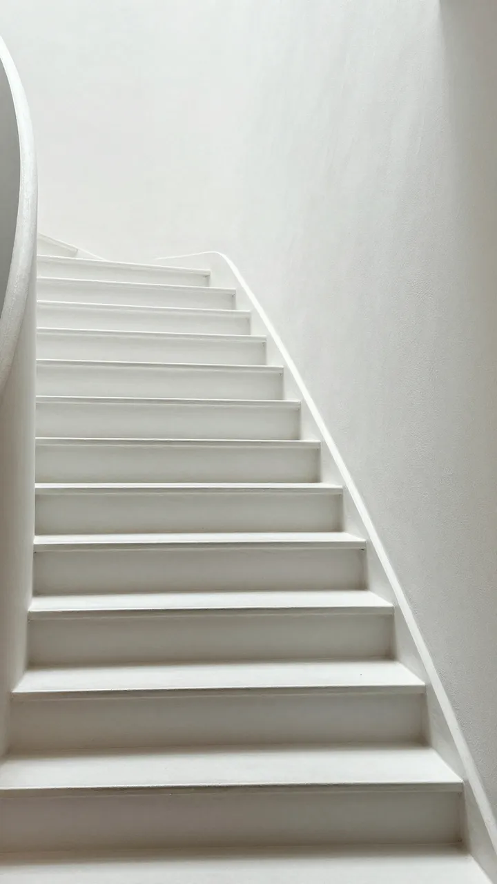 Minimalist white curved staircase ascends alongside smooth plaster walls in soft even light