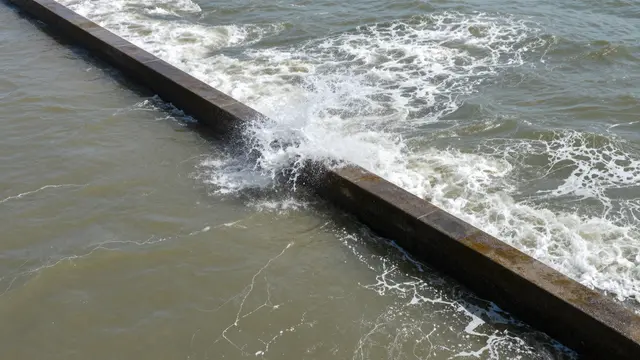 Aerial view of a concrete breakwater with white surf crashing over it during high tide