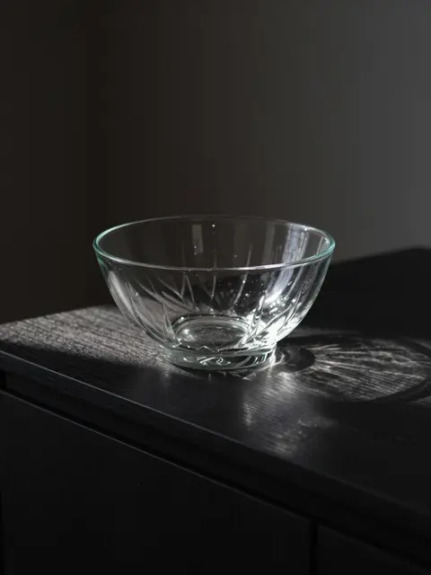 An empty cut-glass bowl catches a single shaft of light on a dark wooden sideboard