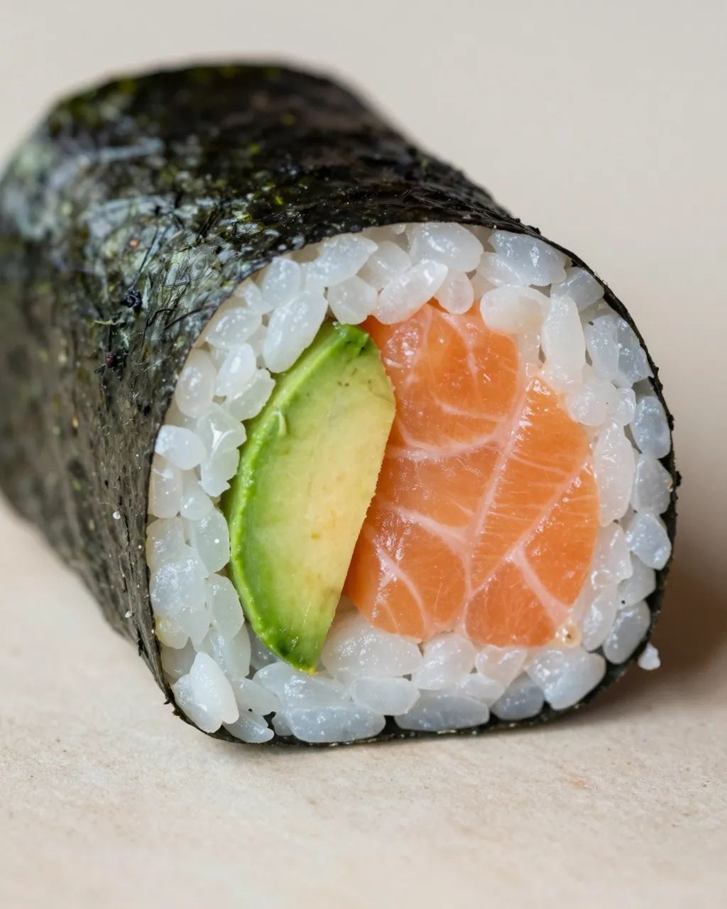 Close-up cross-section of a sushi burrito wrapped in nori, showing rice, raw salmon and avocado filling on a neutral surface.
