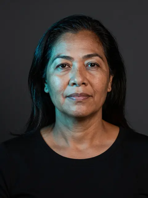 Middle aged Pacific Islander woman in a black top faces the camera with soft cyan lighting on her face against a dark studio