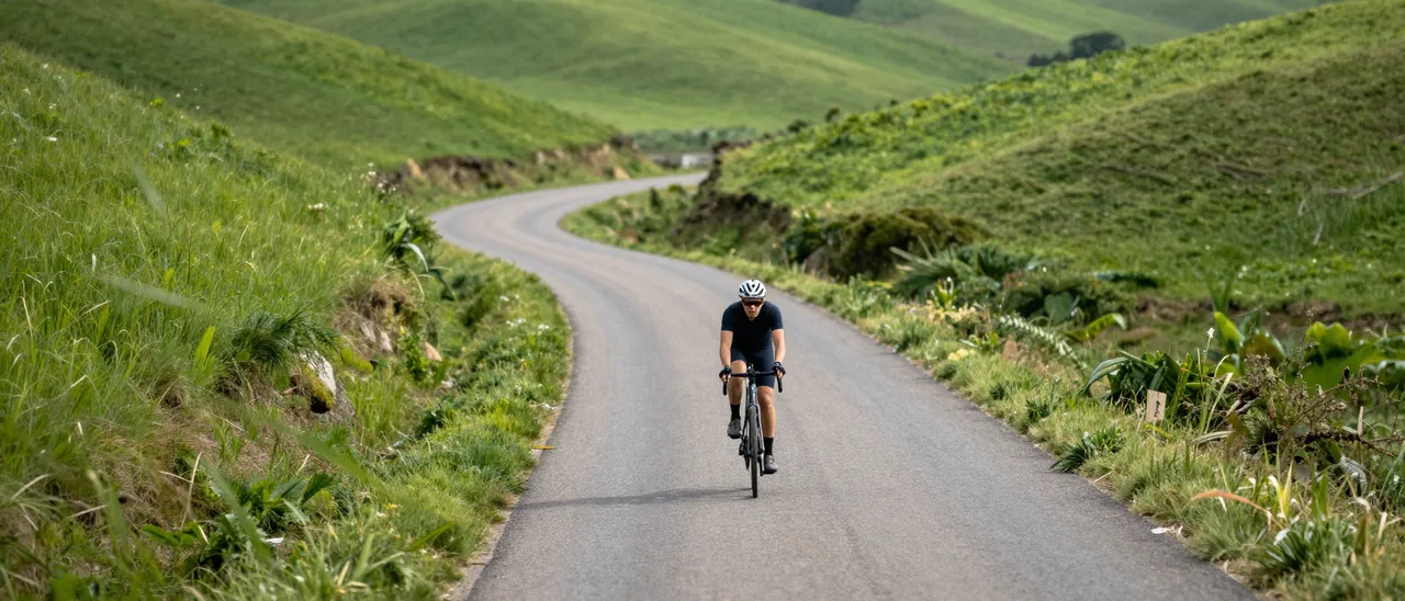 A lone adult cyclist rides a road bike along a winding paved rural road between rolling green hills in soft daylight