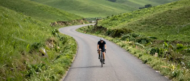 A lone adult cyclist rides a road bike along a winding paved rural road between rolling green hills in soft daylight