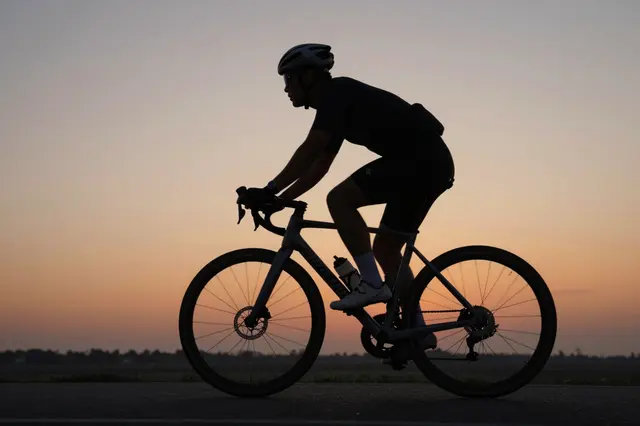 Side profile silhouette of an adult road cyclist riding against a soft orange gradient dusk sky