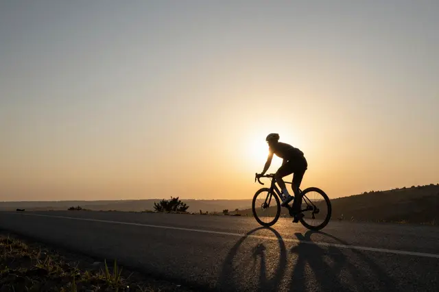 Silhouetted road cyclist rides on a paved hilltop road against a warm setting sun.
