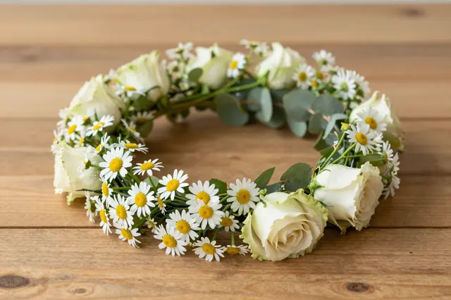 A handmade flower crown of daisies, white roses and eucalyptus rests on a wooden table.