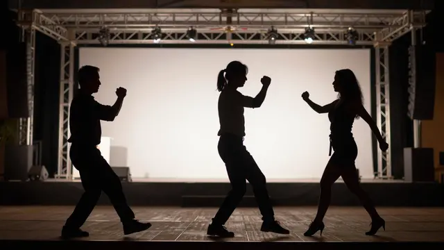 Three adult person silhouettes pose in a synchronized dance in front of a lit outdoor stage at night