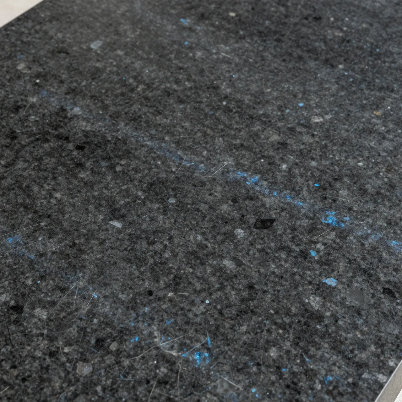 Full frame close-up of polished dark granite speckled with iridescent blue-gray mineral crystals under soft even light.