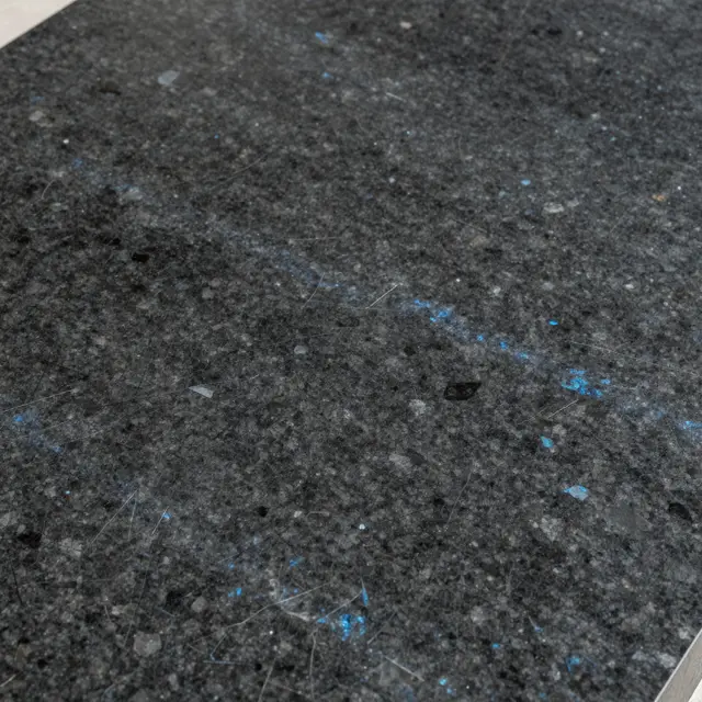 Full frame close-up of polished dark granite speckled with iridescent blue-gray mineral crystals under soft even light.
