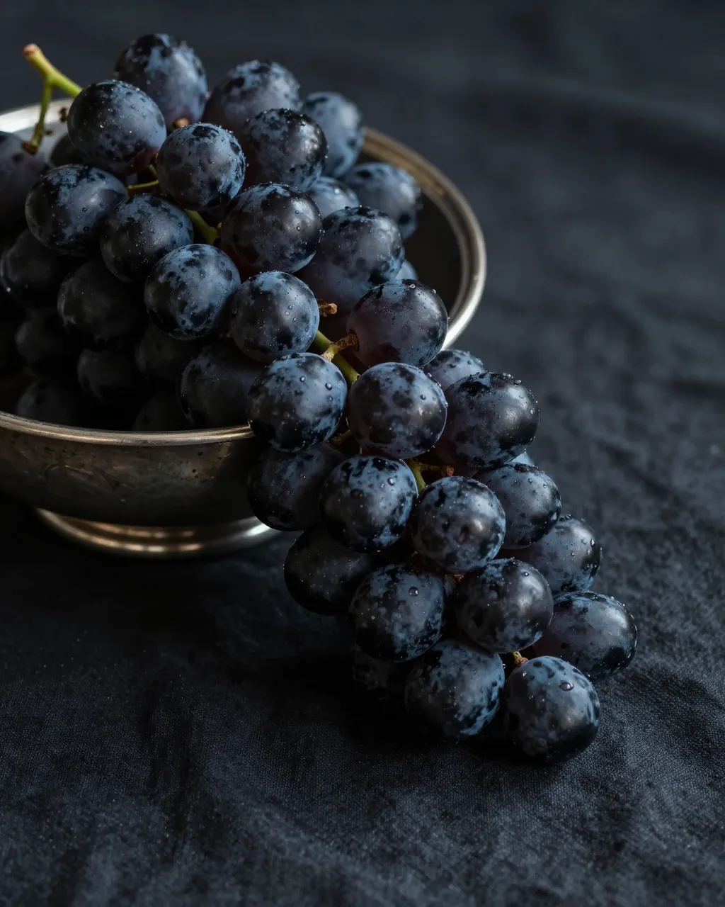 Black grapes spilling from a tarnished silver bowl onto draped dark linen fabric
