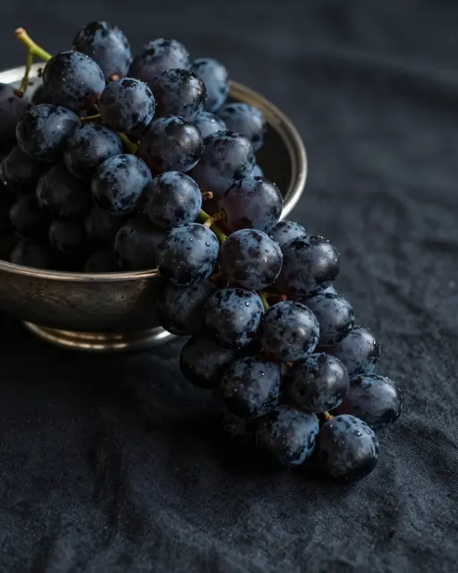 Black grapes spilling from a tarnished silver bowl onto draped dark linen fabric