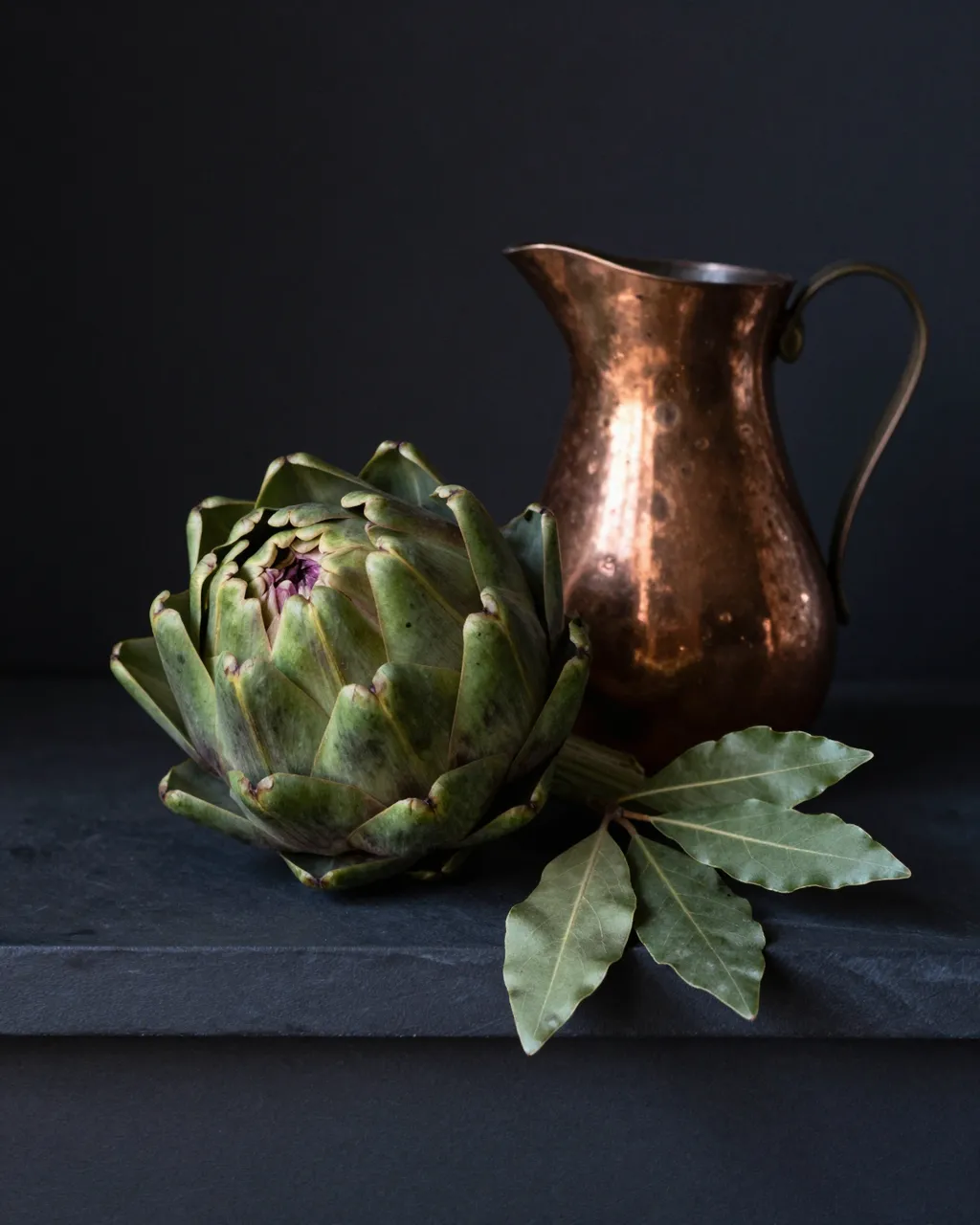 A still life of an artichoke, bay leaves and a hammered copper jug arranged on a dark stone ledge against a black background