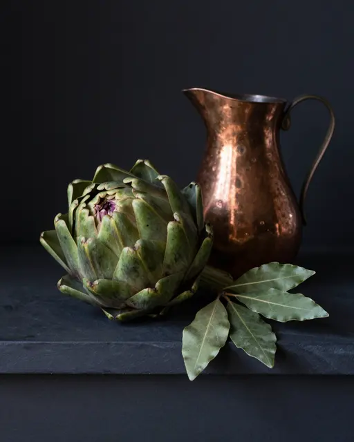 A still life of an artichoke, bay leaves and a hammered copper jug arranged on a dark stone ledge against a black background