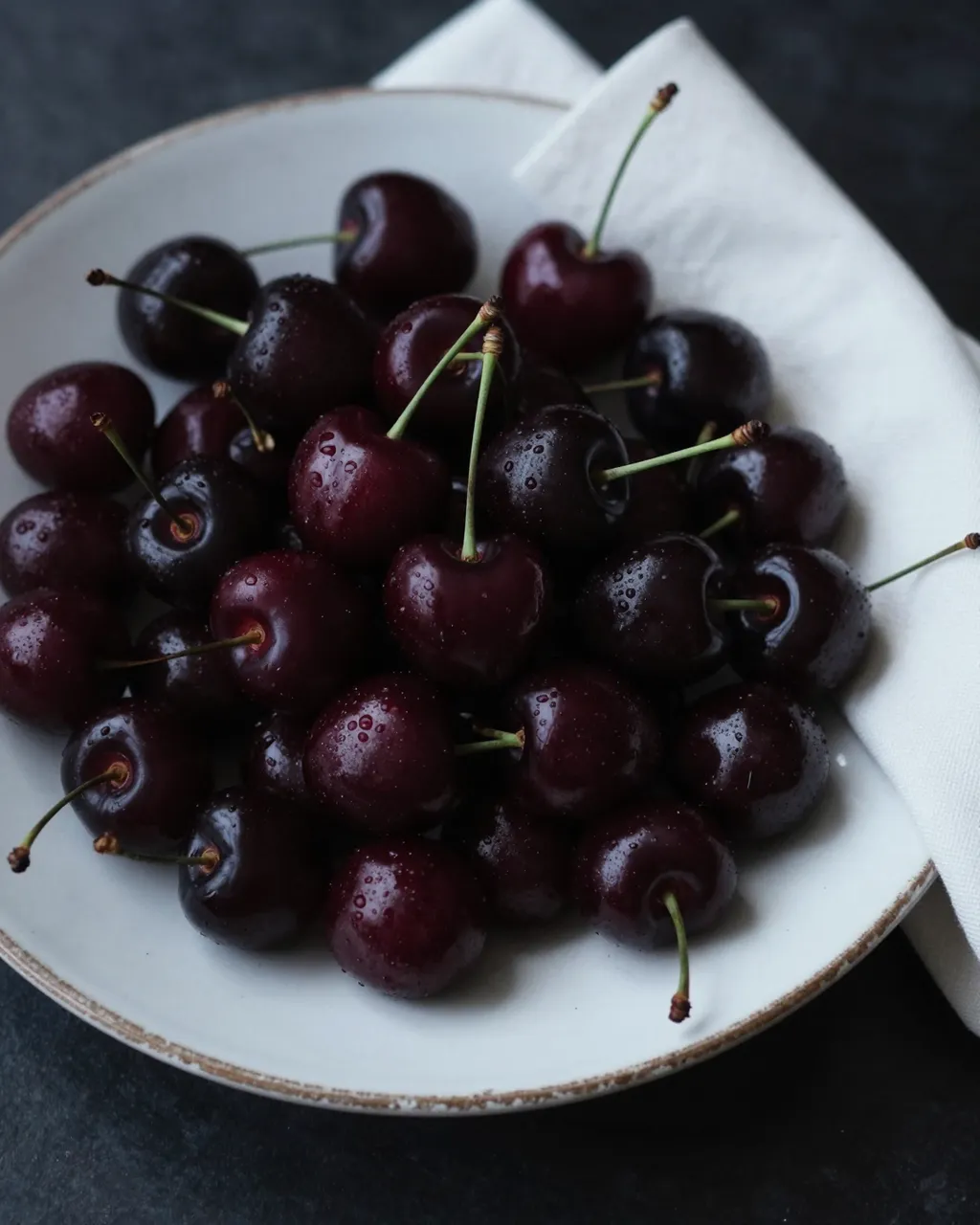 Heap of wet dark cherries resting on a chipped white plate next to a folded white linen napkin over a dark surface