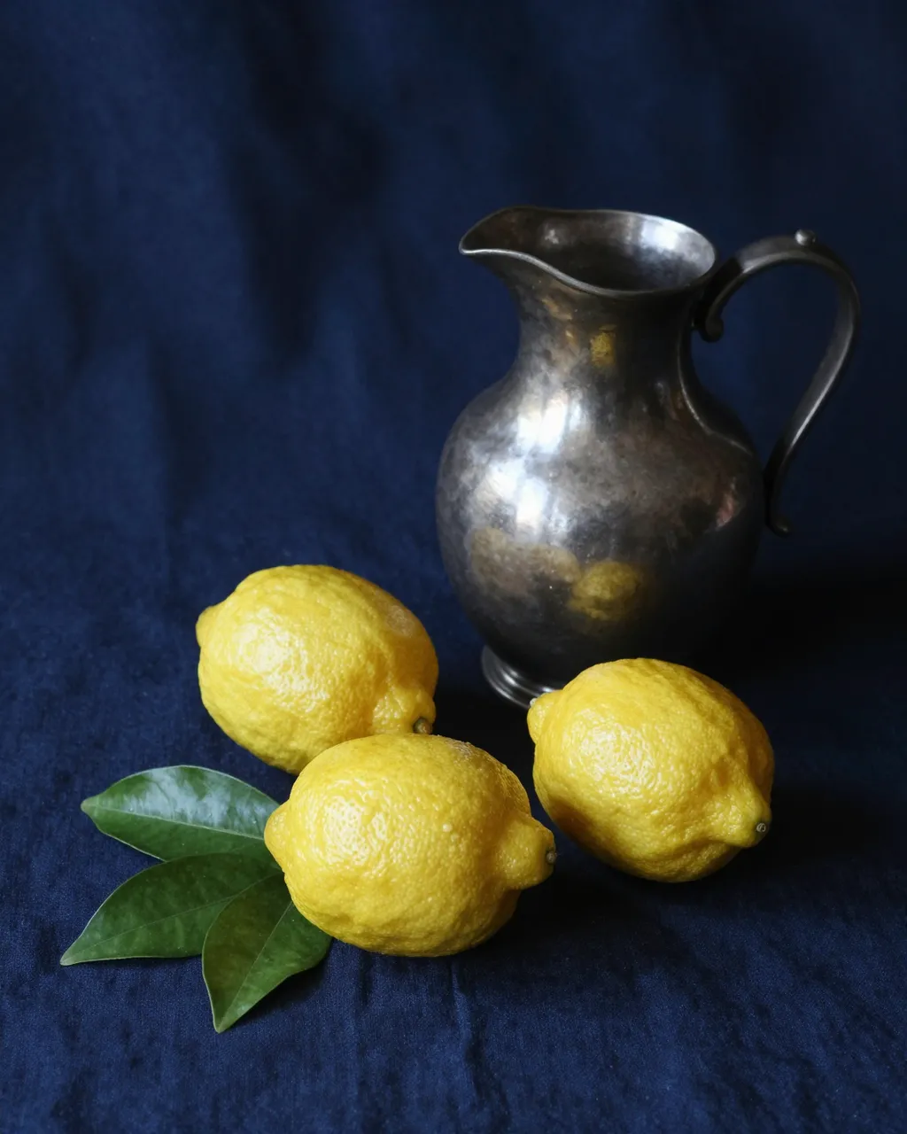 Three lemons with glossy green leaves sit beside a pewter jug on dark blue linen