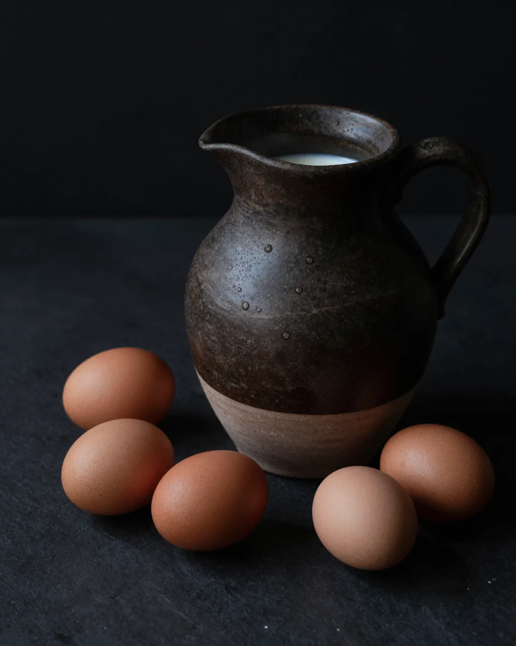 Stoneware pitcher of milk surrounded by brown eggs on a dark matte table against a black background