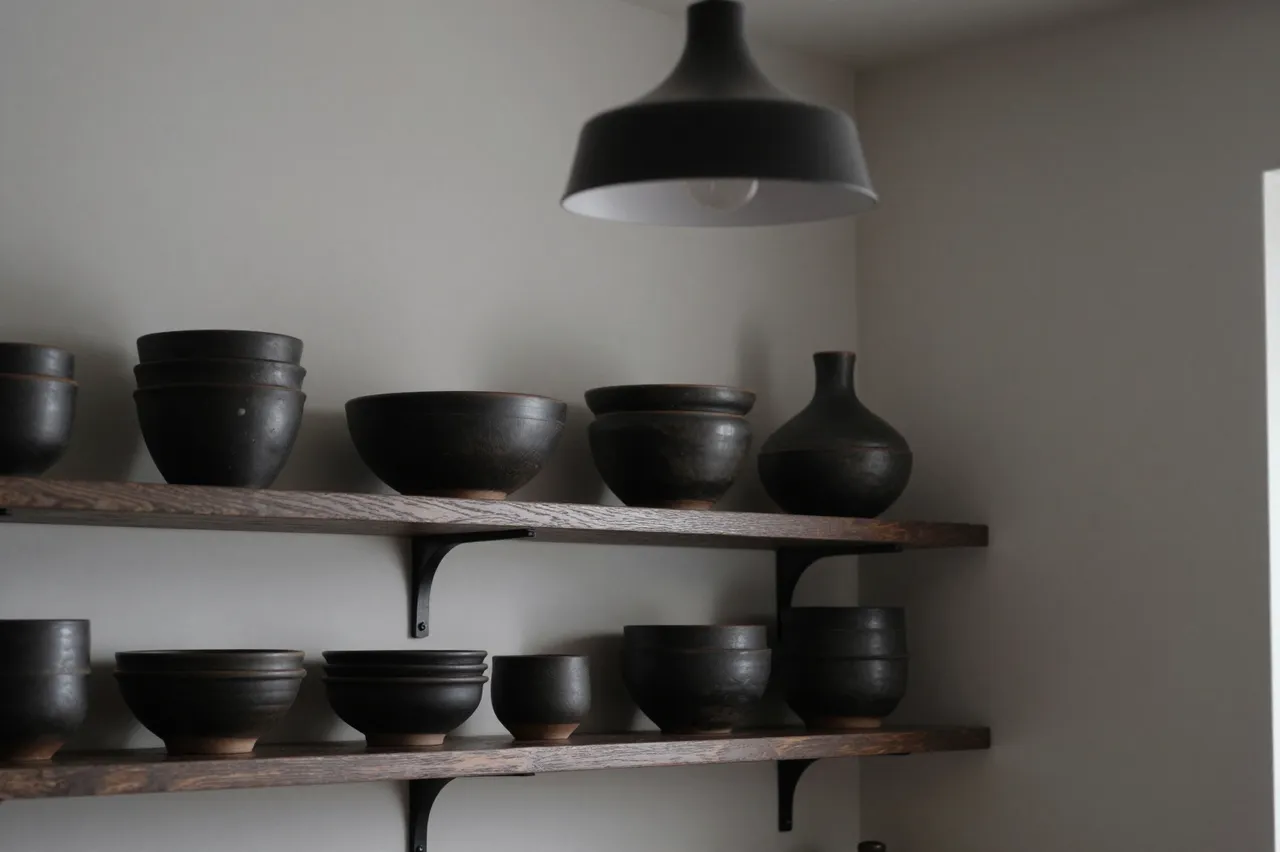 Open wooden kitchen shelves holding dark stoneware pottery under a matte black pendant lamp