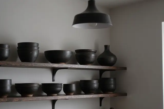 Open wooden kitchen shelves holding dark stoneware pottery under a matte black pendant lamp