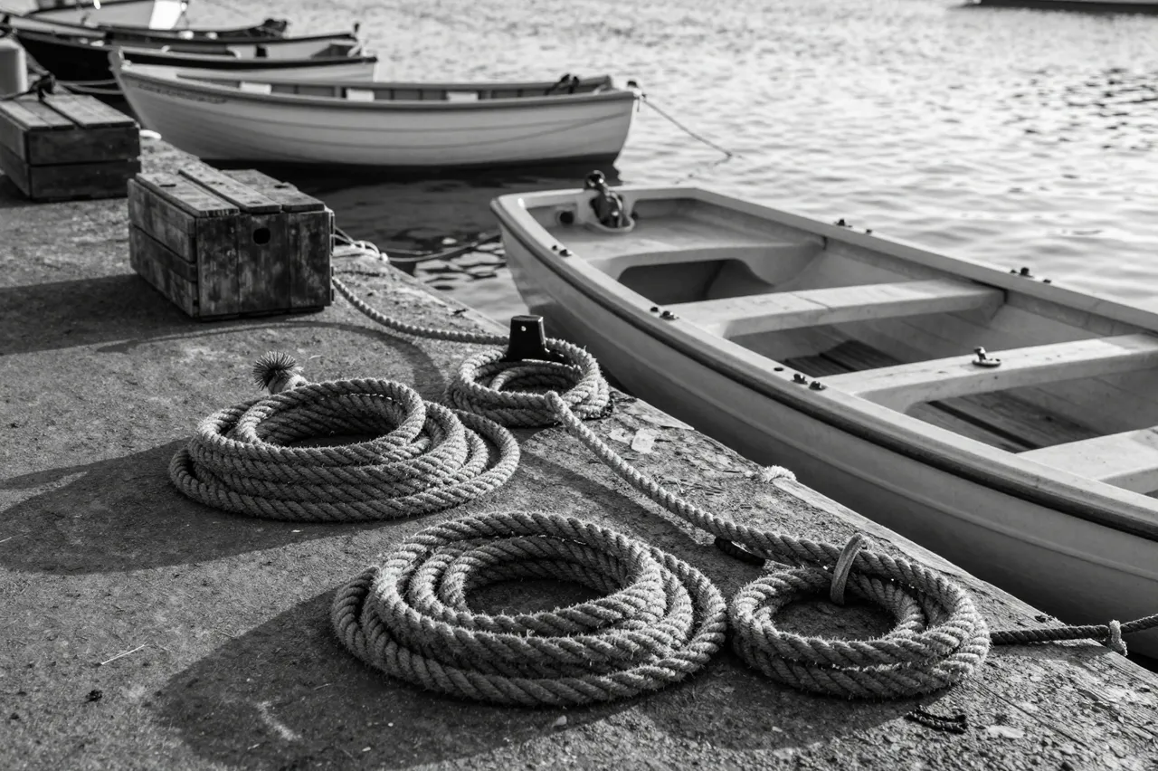 Black and white view of coiled ropes and wooden crates on a harbor quay beside moored rowboats at first light