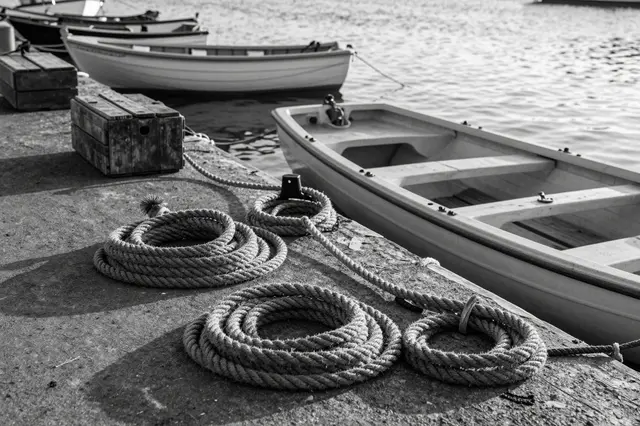 Black and white view of coiled ropes and wooden crates on a harbor quay beside moored rowboats at first light