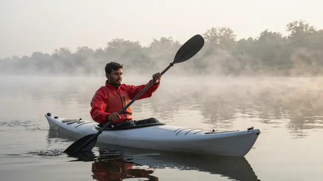 A South Asian man in a red jacket paddles a white kayak on a misty calm river at dawn