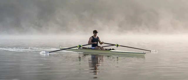 An adult single sculler rows a racing shell across calm misty lake water at sunrise