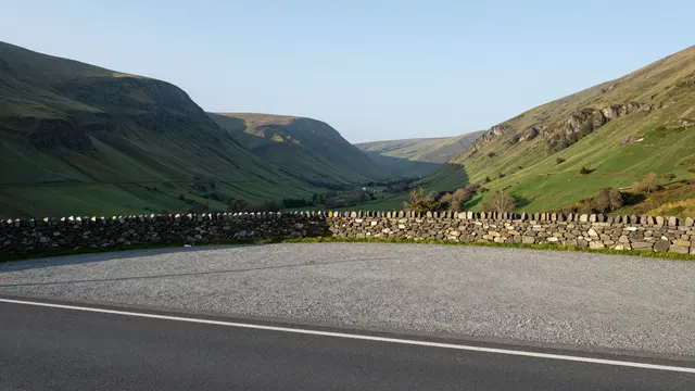 Empty gravel roadside viewpoint with a dry stone wall overlooking a green valley at early dawn