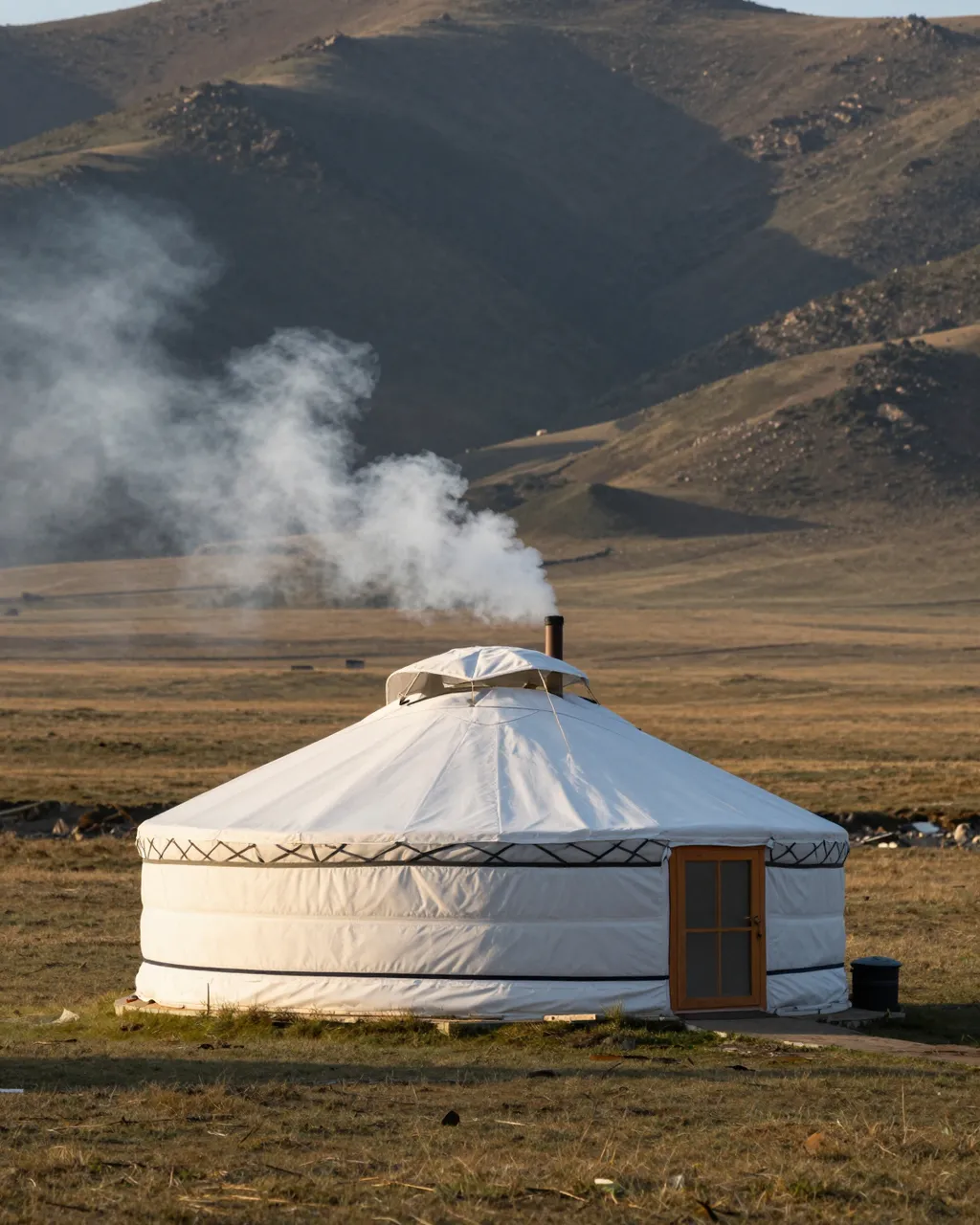 A single white felt yurt with rising chimney smoke sits in a grassy steppe valley at dawn