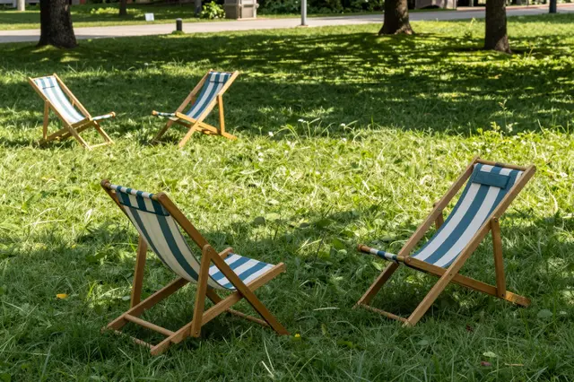 Striped folding deck chairs sit scattered across a sun dappled grassy park lawn under tree shade