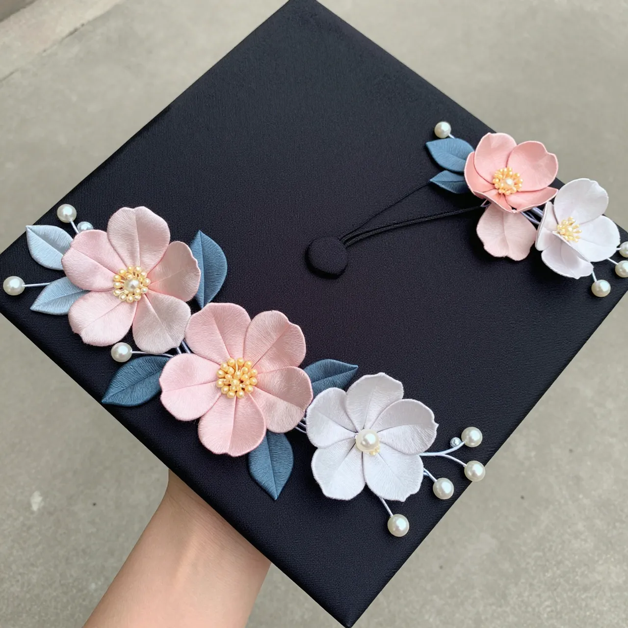 A hand holds a black graduation cap decorated with silk flowers and pearl accents from a top-down perspective.