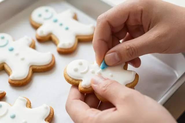 Close-up of hands piping icing onto a gingerbread man cookie over a baking tray