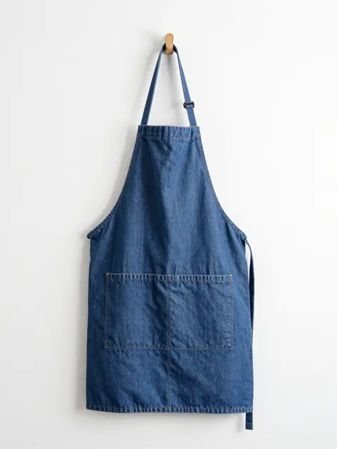 Plain blue denim apron hangs from a wooden peg against a matte white wall