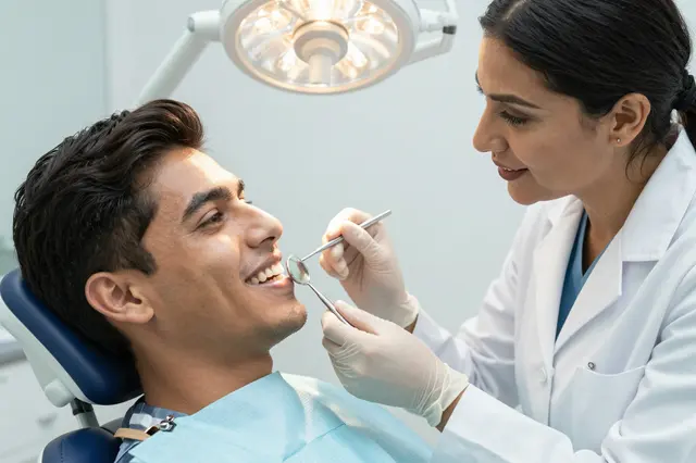 A female South Asian dentist examines a smiling male patient's teeth in a bright dental clinic.
