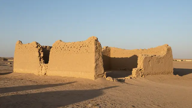 Mud-brick caravan inn ruins stand on a sandy desert plain under clear sky at sunset with long shadows