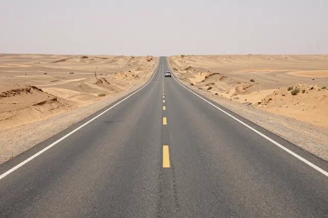 A straight paved desert road stretches to the horizon with a small car traveling far ahead