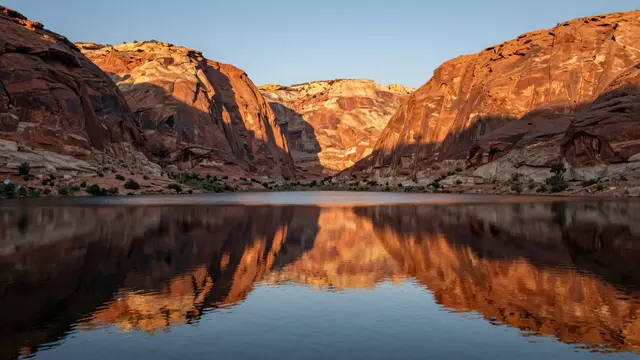 Calm desert lake reflects sunlit red rock canyon cliffs at golden sunset