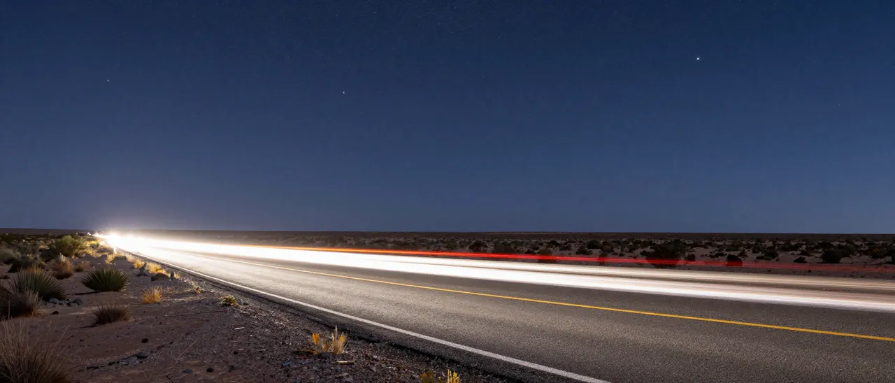 Long exposure light trails from passing vehicles stretch across a desert road under a dark starry night sky