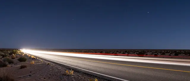 Long exposure light trails from passing vehicles stretch across a desert road under a dark starry night sky