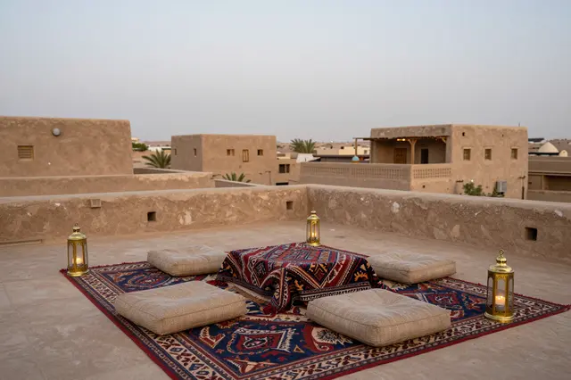 An earthen rooftop terrace set with woven rugs, floor cushions and brass lanterns overlooks an adobe desert town at dusk