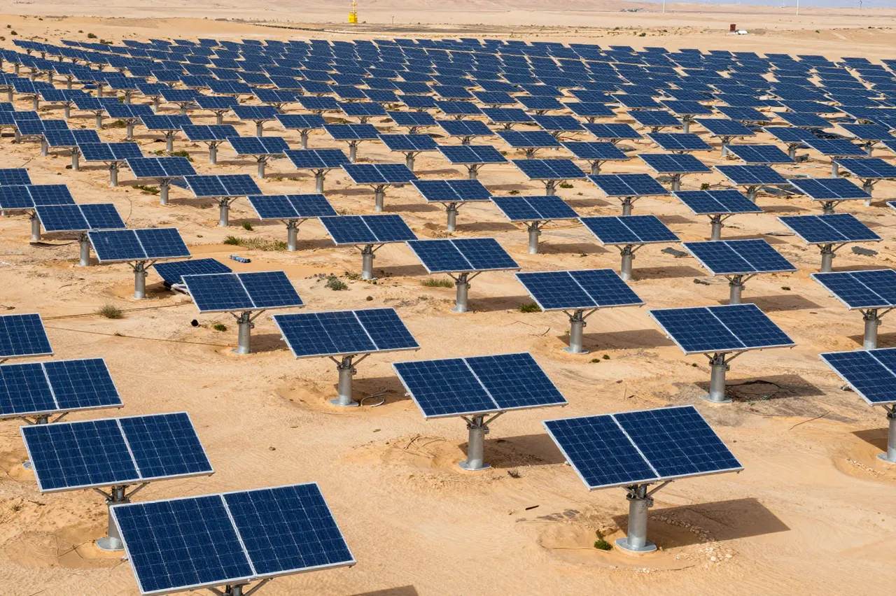 Rows of photovoltaic solar panels stretch across an arid sandy desert landscape under bright daylight