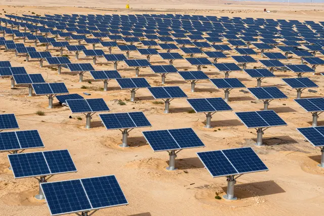 Rows of photovoltaic solar panels stretch across an arid sandy desert landscape under bright daylight