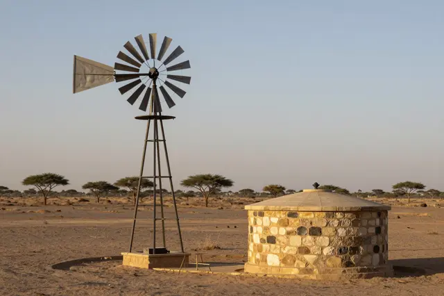 Metal windpump beside a stone water tank on an arid plain with scattered acacia trees at evening