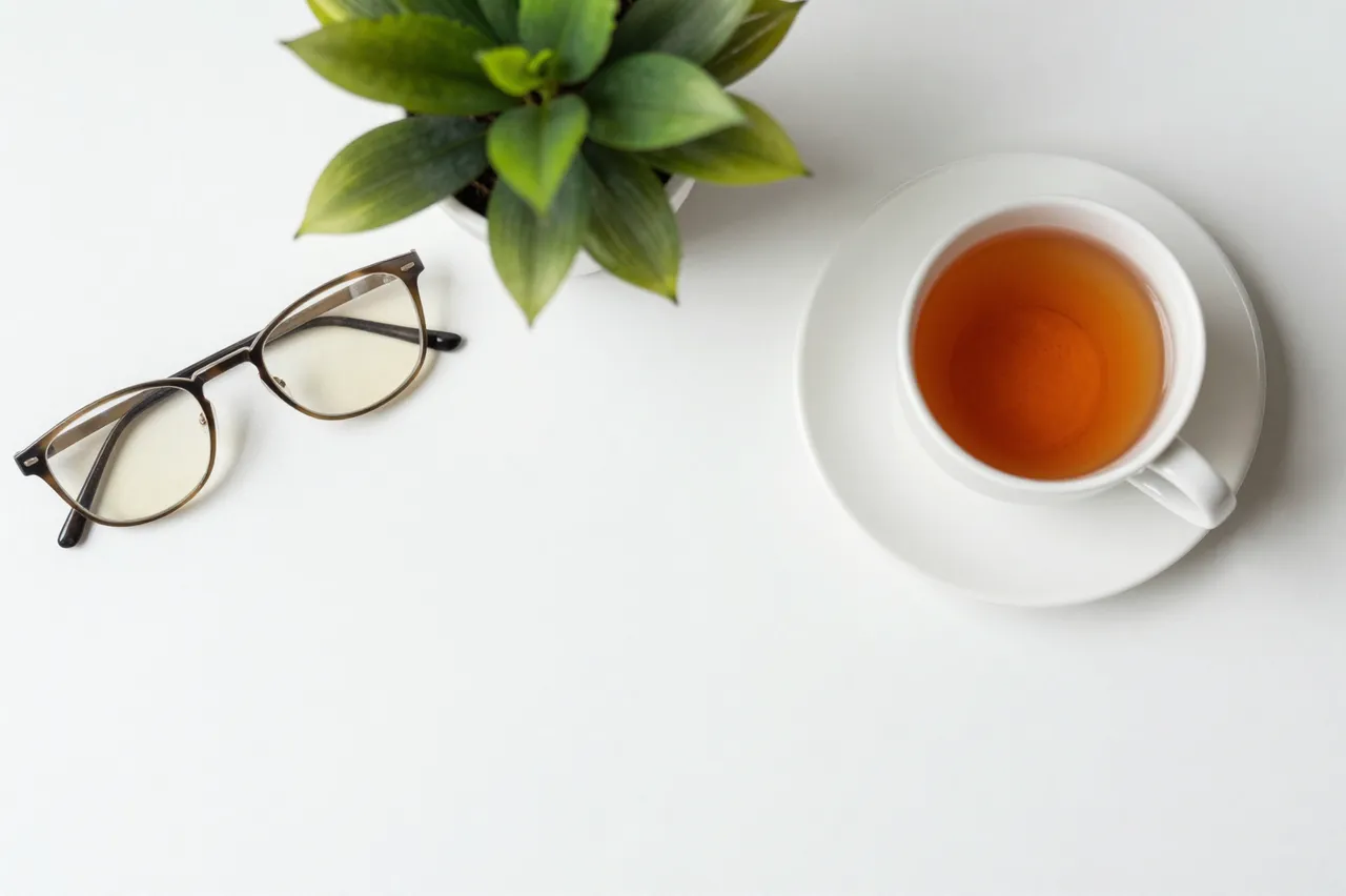 Top-down view of a white desk holding eyeglasses, a potted plant, and a cup of tea with empty copy space.