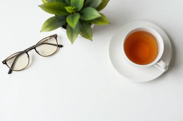 Top-down view of a white desk holding eyeglasses, a potted plant, and a cup of tea with empty copy space.