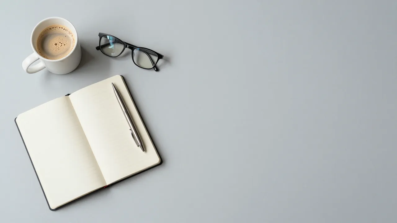 Top down view of a pale gray desk with an open notebook, pen, latte mug, and glasses on the left, empty space on the right.