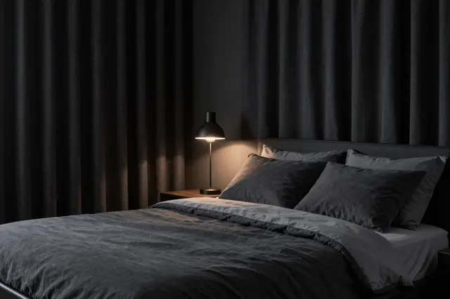 A tidy dark bedroom with a made bed, blackout curtains and a glowing small bedside lamp