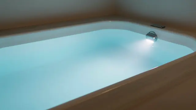 A shallow still pale blue lit pool sits in a dim enclosed float therapy room